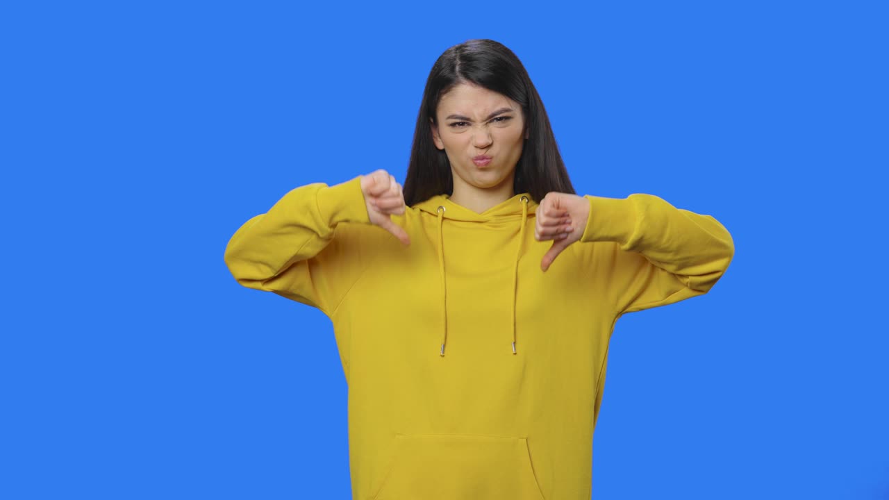 Portrait of pretty brunette showing thumbs down gesture. Young woman with long hair in yellow sweatshirt posing in studio with blue screen background. Close up. Slow motion ready 59.94fps