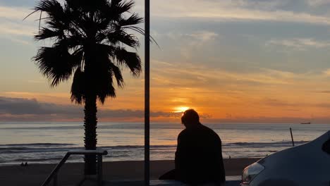 silueta de un joven adulto fumando un cigarrillo al atardecer y con algunos surfistas disfrutando del mar