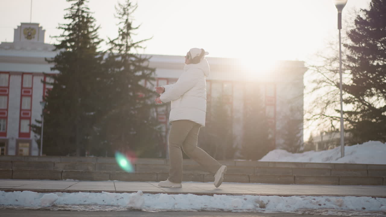 Dancer in white winter coat moves gracefully across step pavement, surrounded by snow piles and tall trees, with warm sunset casting silhouette and flare through background architecture