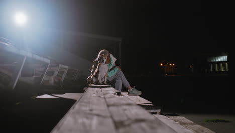 coach sitting with puppy on plastic bench under night sky after training session, illuminated by bright spotlight, enjoying peaceful quiet moment in outdoor park surrounded by distant building lights