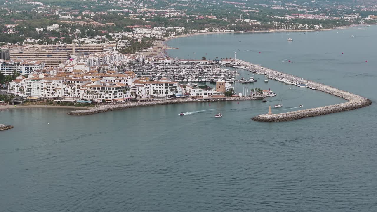 Aerial view of Marbella Marina with whitewashed buildings, boats, and breakwaters extending into the Mediterranean Sea, backed by rolling hills