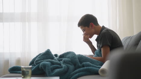 ill boy sneezing into tissue seated on couch with blanket covering him near window, showing tiredness and fatigue, feeling unwell and needing rest while managing cold symptoms
