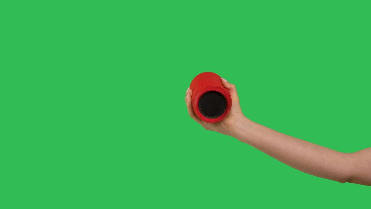 Cropped shot of female hand holding a wireless red bluetooth speaker on green screen chroma key background. Musical electronic gadget for listening to music. Close up. Slow motion