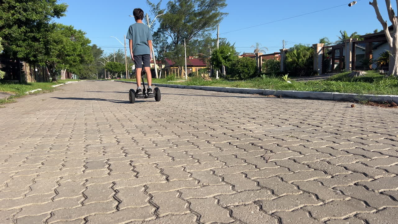 Teen boy steps onto hoverboard and rides away down residential street, starting with board alone in frame, low angle wide shot, 4K30