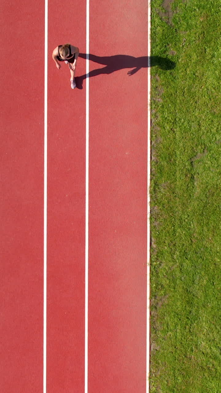 A vertical aerial shot of a fitness girl jogging on a running track next to green grass. Perfect for sports, health, and motivation content