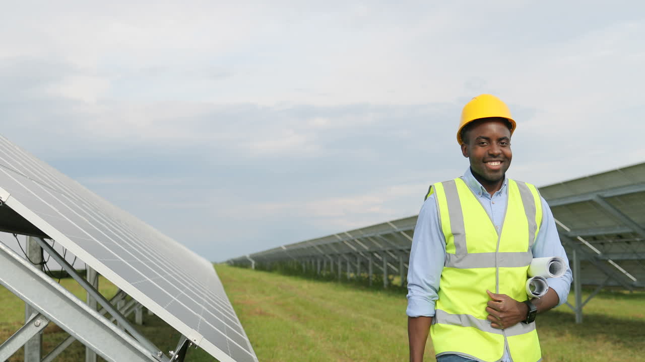 Happy african american engineer walking and holding rolls of paper with diagrams and tool box on solar plantation