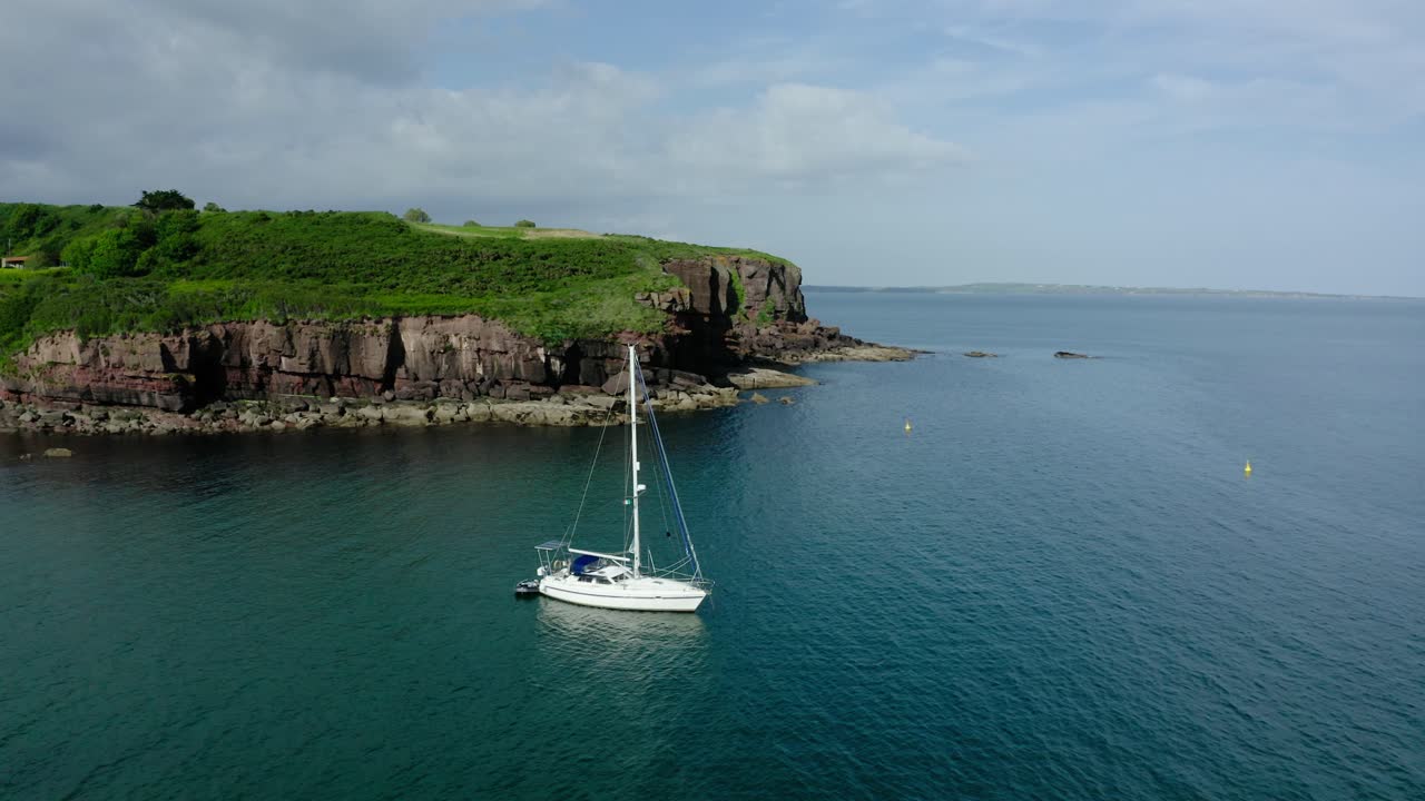 A sailboat anchored off the coast of Ireland with steep cliffs in the background.