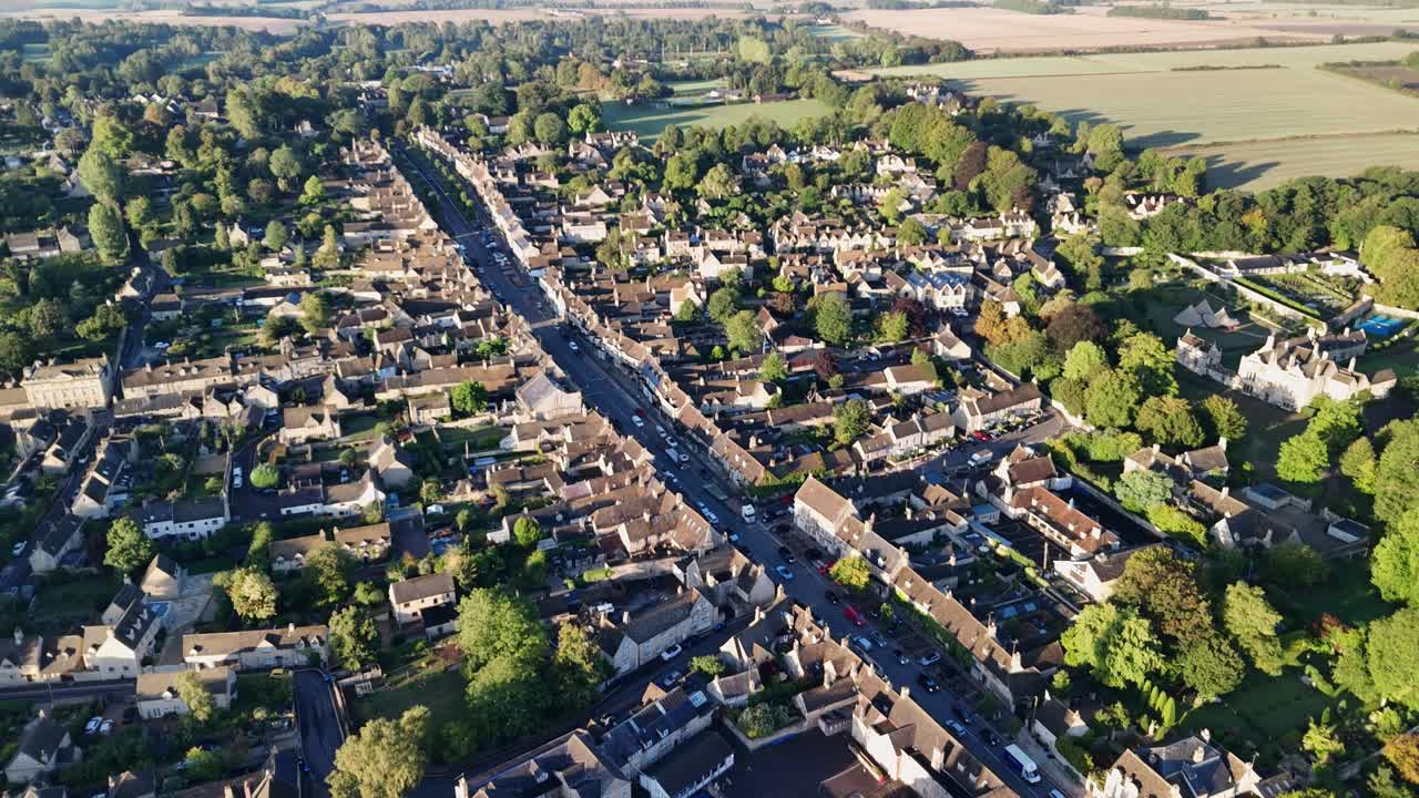 aerial drone flyover of the village of Burford in the Cotswolds, the historic High Street and golden stone buildings, bathed in the early morning sunlight
