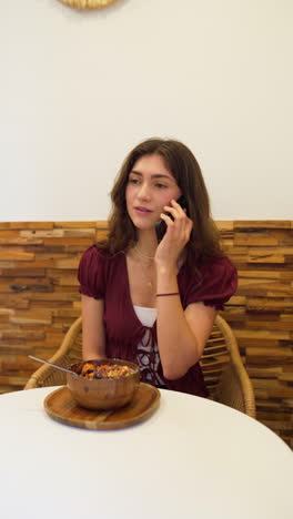 young woman enjoying a delicious, healthy acai bowl at a trendy cafe, happily chatting on her phone while savoring the moment.