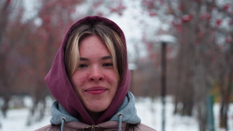 primer plano de una mujer joven con una chaqueta con capucha cálida, sonriendo con las manos en los bolsillos mientras está de pie en un camino nevado, figura borrosa visible en la distancia, árboles de bayas rojas y farolas