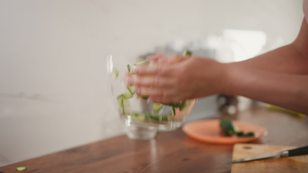 Hand view of person shaking sliced cucumber in transparent glass bowl on kitchen table, with some pieces falling out during preparation, capturing real kitchen activity in soft light
