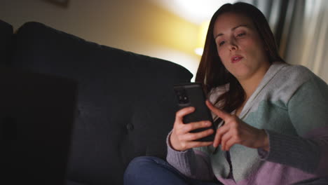 mujer sentada en el sofá en casa por la noche transmitiendo o viendo una película o un programa en la computadora portátil y desplazándose por internet en el teléfono 13