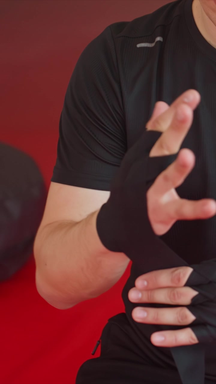 Close up of man in black wrapping his right hand. Visible skin texture hair on arm as man methodically wraps. Red floor suggests indoor fitness space like gym or home workout