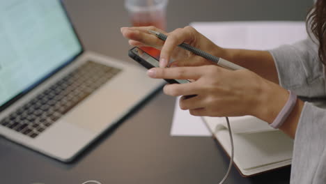 Joven-Mujer-De-Negocios-Asiática-Escribiendo-Notas-Usando-Un-Teléfono-Inteligente-Revisando-Mensajes-Disfrutando-De-Estudiar-Escuchando-Música-Usando-Auriculares-Estudiante-En-El-Lugar-De-Trabajo-De-Oficina-De-Cerca