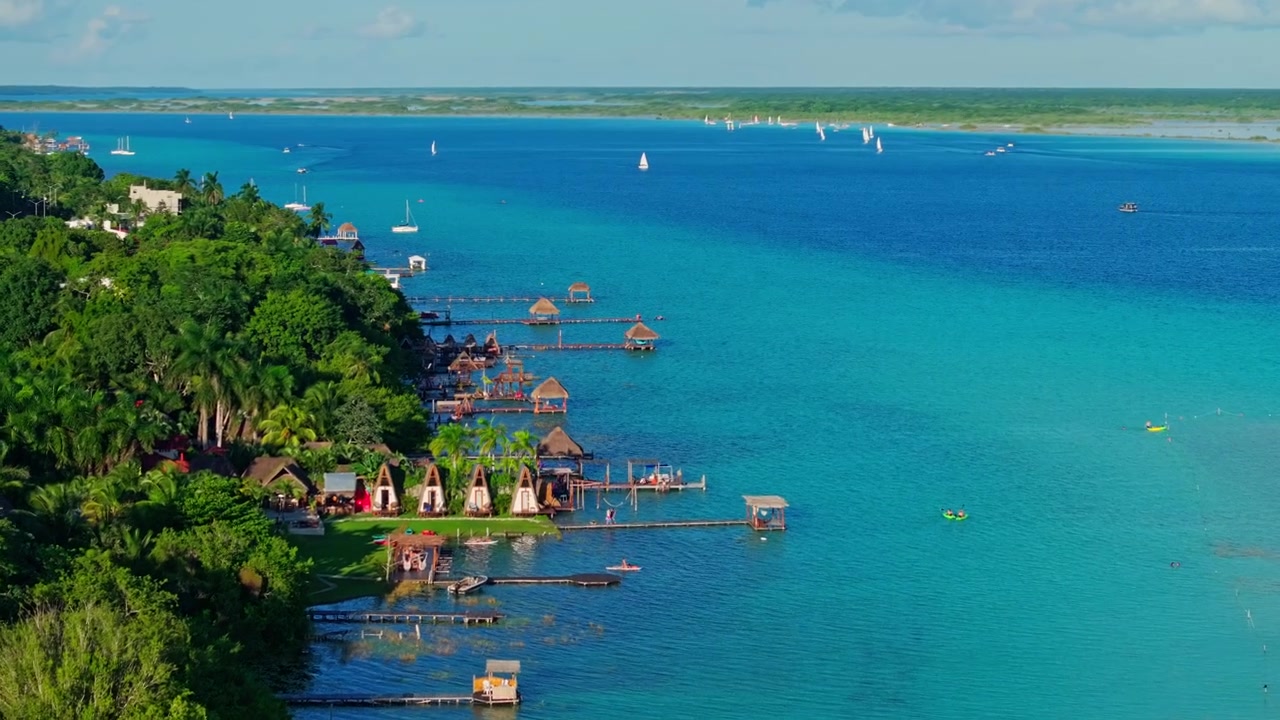 Luftfoto af træmoler og palapaer langs søbredden af Bacalar-lagunen, Mexico, med anløbsbroer, der strækker sig ud over det levende turkisblå vand.