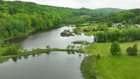 Una-Pintoresca-Toma-Aérea-De-Una-Zona-Turística-Cerca-De-Un-Lago-Con-Cabañas,-Césped-Verde-Y-Colinas-Boscosas-Circundantes