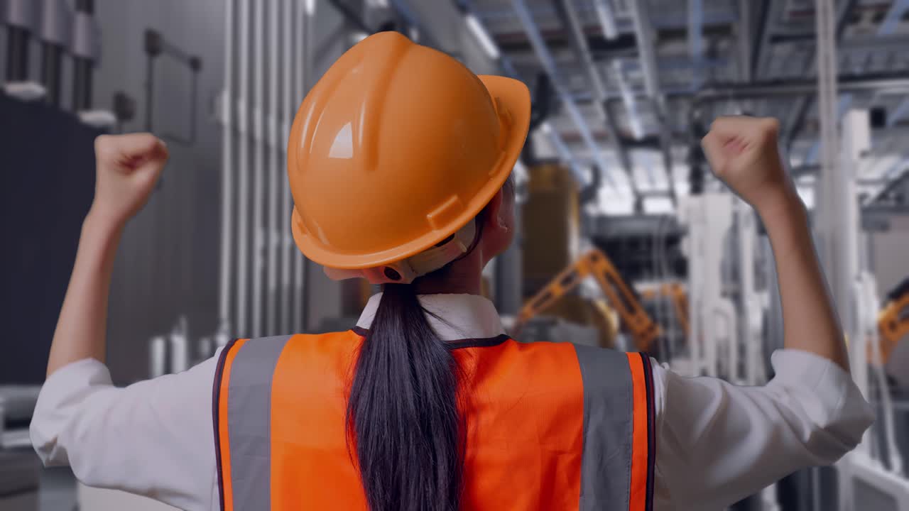 Primo piano da dietro di un'ingegnera con elmetto di sicurezza che alza le mani per festeggiare mentre lavora, linea di produzione automatizzata di batterie in una fabbrica moderna.