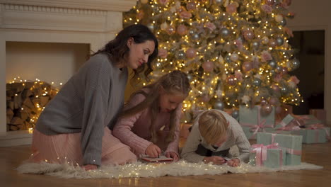 niño y niña escriben una carta con su madre en el suelo junto a los regalos y el árbol de navidad