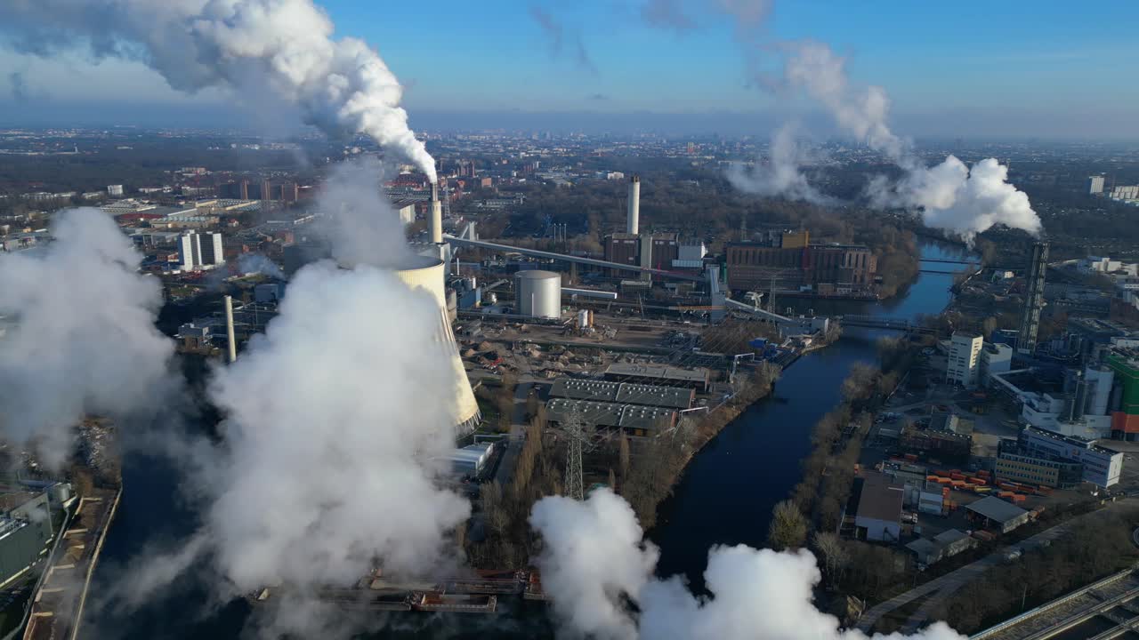 Aerial view of a thermal power plant emitting smoke on a sunny day, with a river and a city in the background. Nice aerial view flight speed ramp hyper motion time lapse
