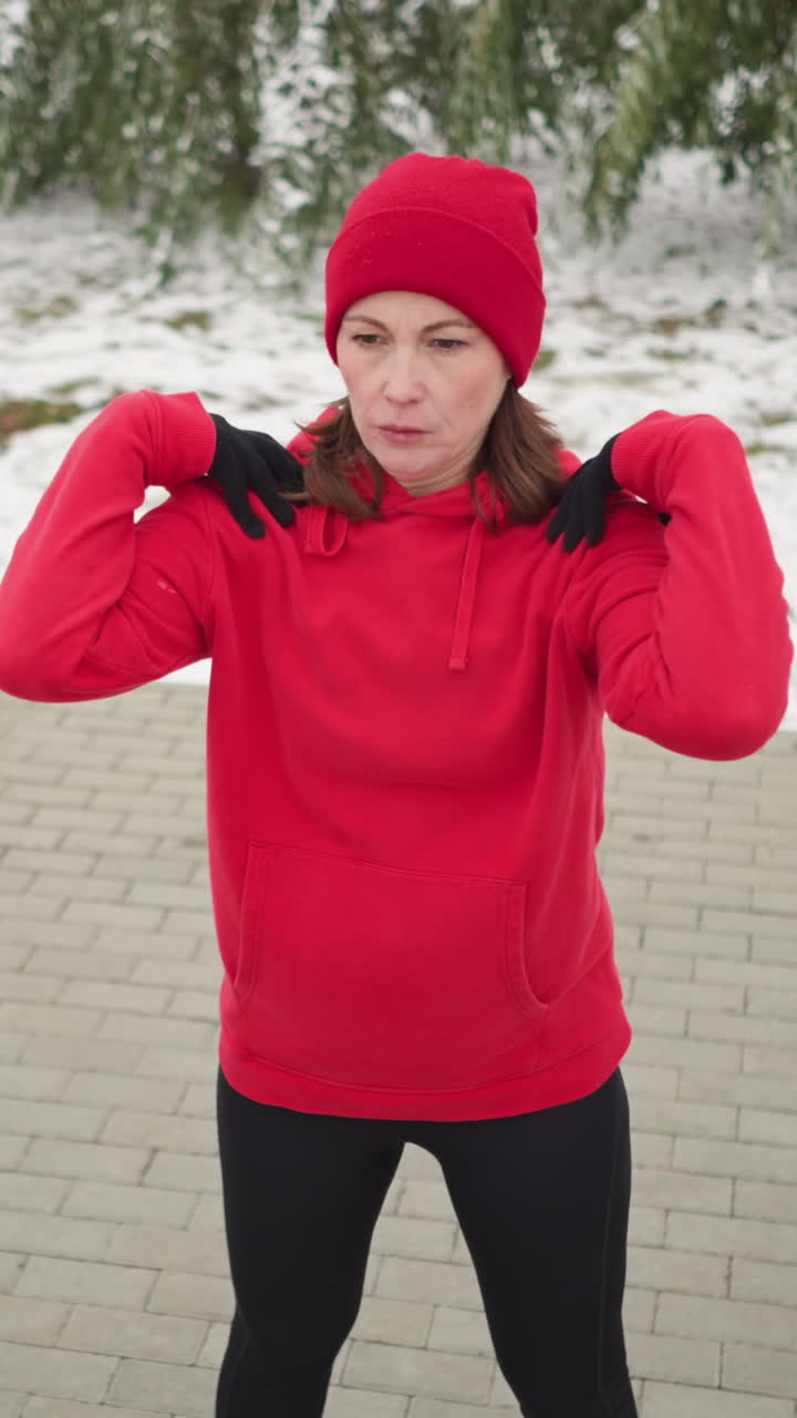 woman performing hand stretch turning her arm in outdoor fitness routine, dressed in athletic wear, background features frosted tree, bag on bench, snow-covered ground, and serene winter setting