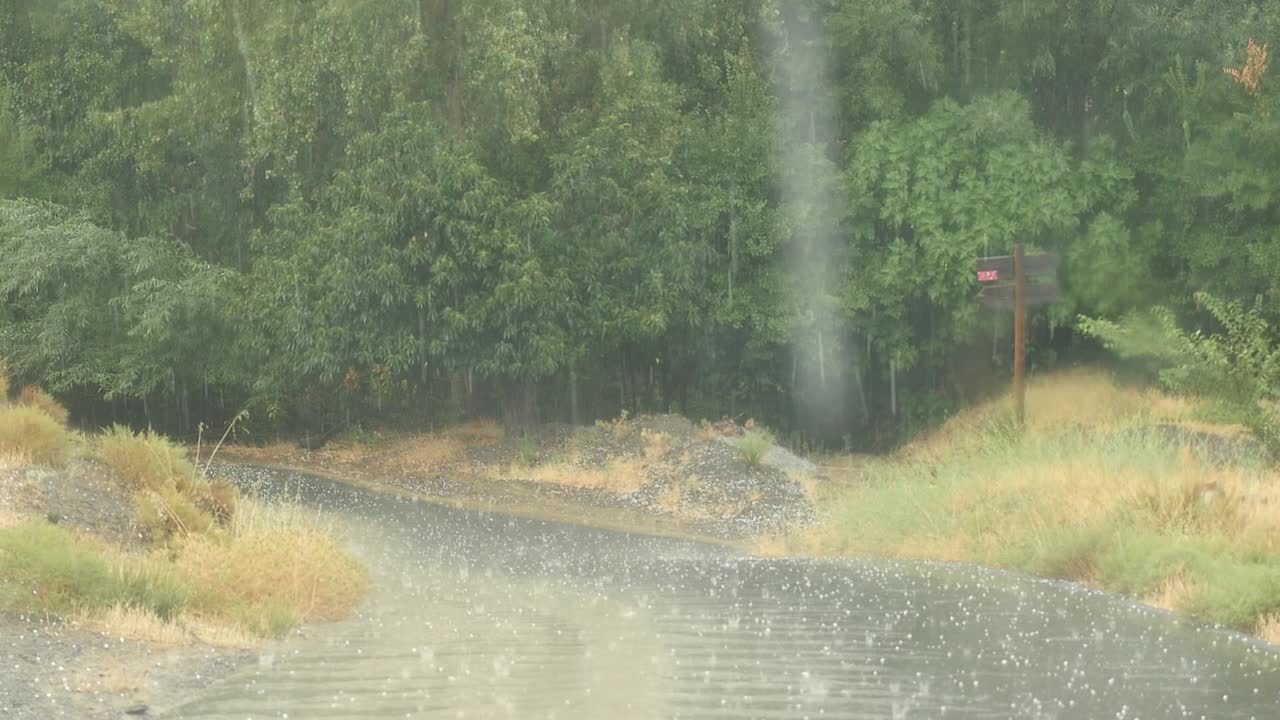 Severe rain and hail storm on a rural road, captured from inside a car. Raindrops are visible on the windshield, as is the movement of the windshield wipers