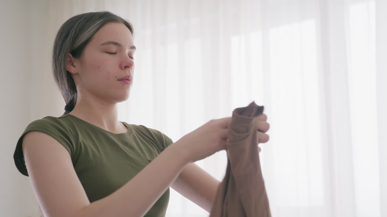 Lady carefully folding brown polo shirt in bright living room with soft daylight filtering through curtains, representing household chores, tidiness, organisation, and domestic lifestyle routine