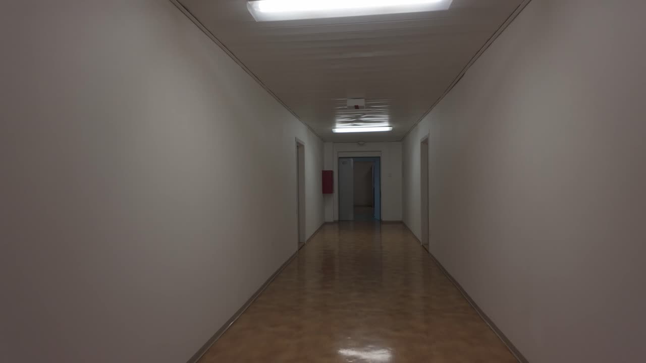 A hotel corridor leading straight past the rooms, lit by fluorescent lights. Walking from a first-person perspective