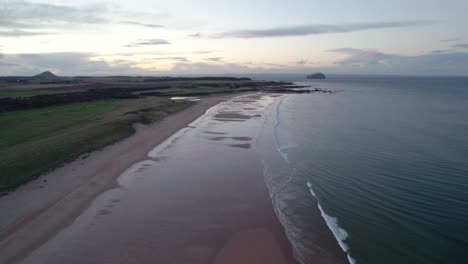 Drohnenaufnahmen-Hoch-über-Einem-Langen-Sandstrand-Bei-Sonnenuntergang-Mit-Blick-Auf-Den-Plätschernden-Ozean,-Während-Die-Flut-Sanft-Die-Küste-Umspült