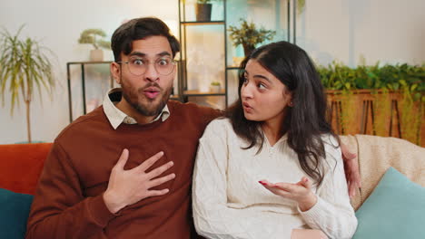 amazed shocked indian couple with wow reaction open mouth looking at each other sitting on home sofa