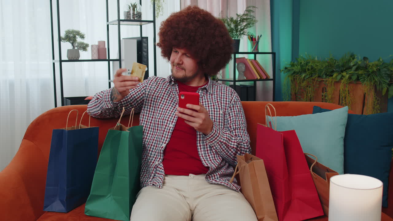 joven adicto a las compras feliz sentado con bolsas de compras haciendo pagos en línea con tarjeta de crédito en casa