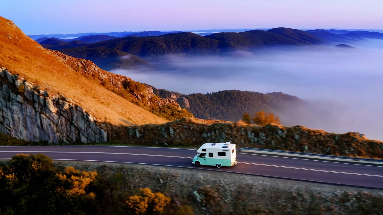 White and mint camper van moves on curving paved highway across rocky golden slopes, overlooking forested ridgelines, mist filled valleys and soft dawn light over wide scenic terrain