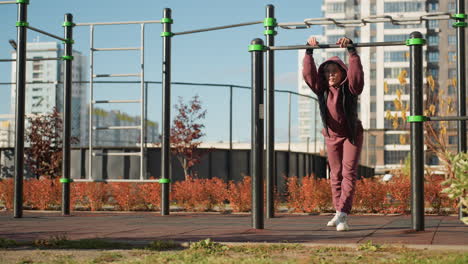wellness minded woman gripping metal bar with both hands leaning forward under bright sun hood up intense gaze on determined face urban park pull up exercise showing strength resilience