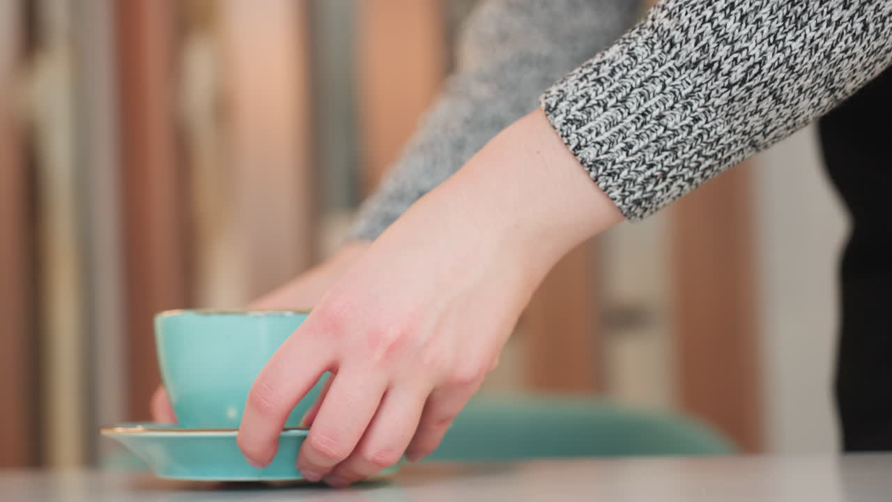 Elegant young woman gently walking toward table bending slightly cold drink held in both hands carefully placing cup on table preparing to sit cozy indoor setting warm ambiance soft lighting