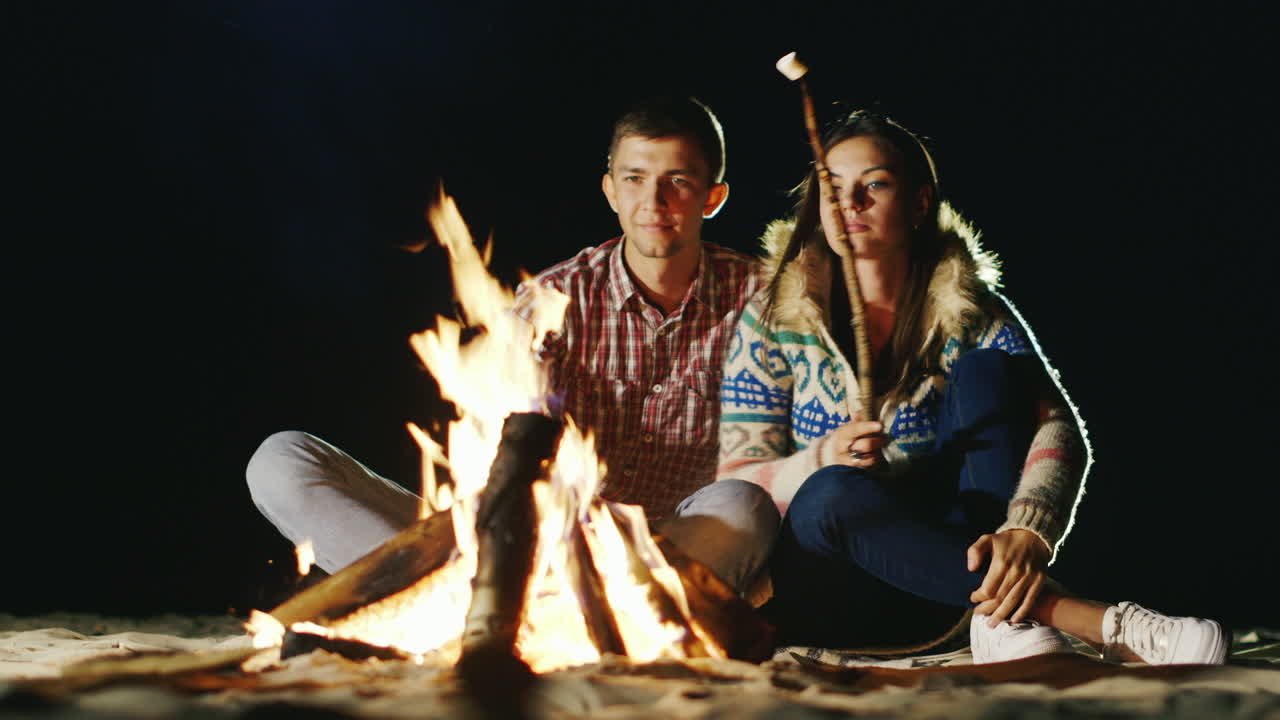 мужчина и женщина отдыхают вечером у костра, жарят зефир на палочках для костра