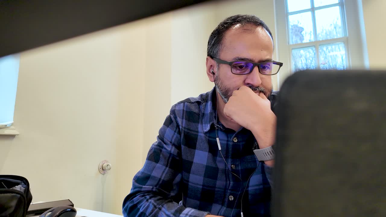 Uomo dell'Asia meridionale con occhiali per luce blu concentrato al computer portatile in ufficio, indossa una camicia a quadri. Lavora da remoto da casa: concetto di lavoro e tecnologia.