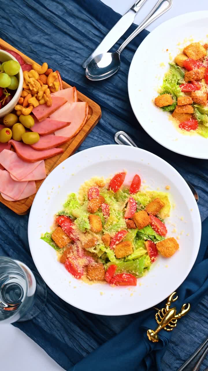 Insalata Caesar e Tagliere di Snack su una Tavola Apparecchiata con Lino Blu. Vista dall'Alto dell'Insalata con Crostini e del Tagliere con Prosciutto e Uva