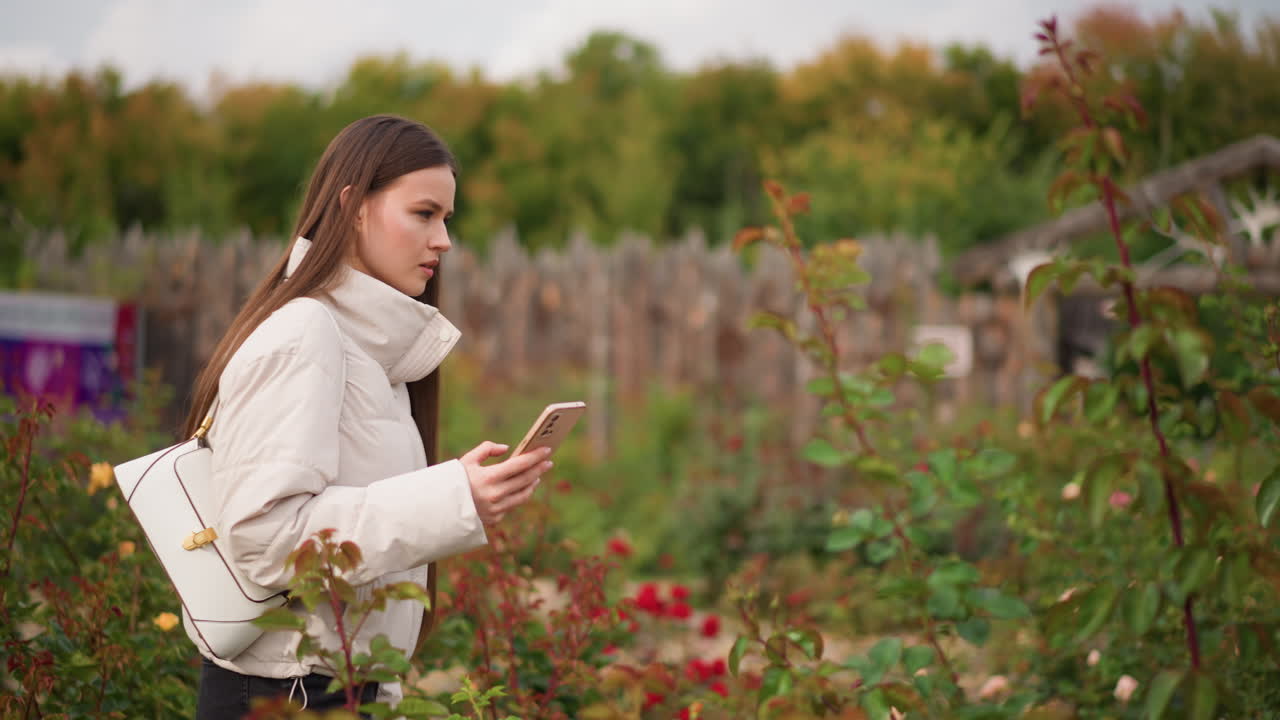 Giovane donna che passeggia in un giardino con il telefono, controlla i messaggi, si ferma a fotografare le rose, esplora i cartelli dell’orto botanico, indossa una giacca trapuntata e una sciarpa, porta una borsa a tracolla, fogliame autunnale