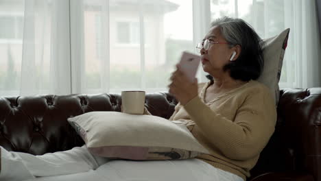 active senior woman relaxing at home wearing bluetooth earbud enjoy listening to music from a mobile phone sitting comfortably on a sofa