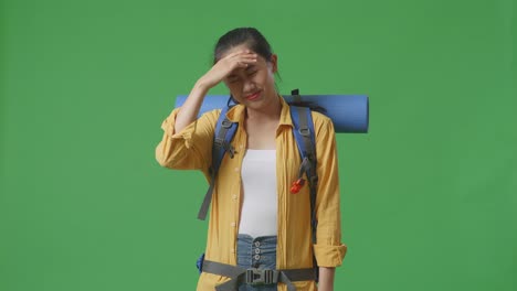asian female hiker with mountaineering backpack having a headache while standing on green screen background in the studio