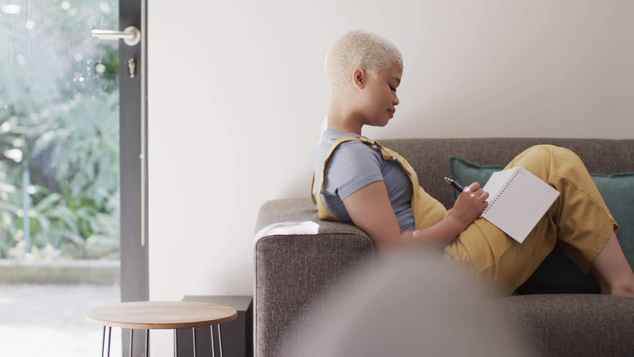 Happy biracial woman taking notes and sitting on couch in slow motion