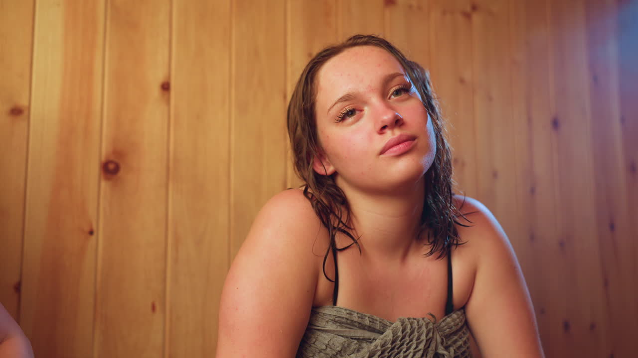 woman seated on wooden bench inside sauna relaxes and interacts with unseen companion in background, damp hair resting on shoulders, warm amber light accentuating serene mood