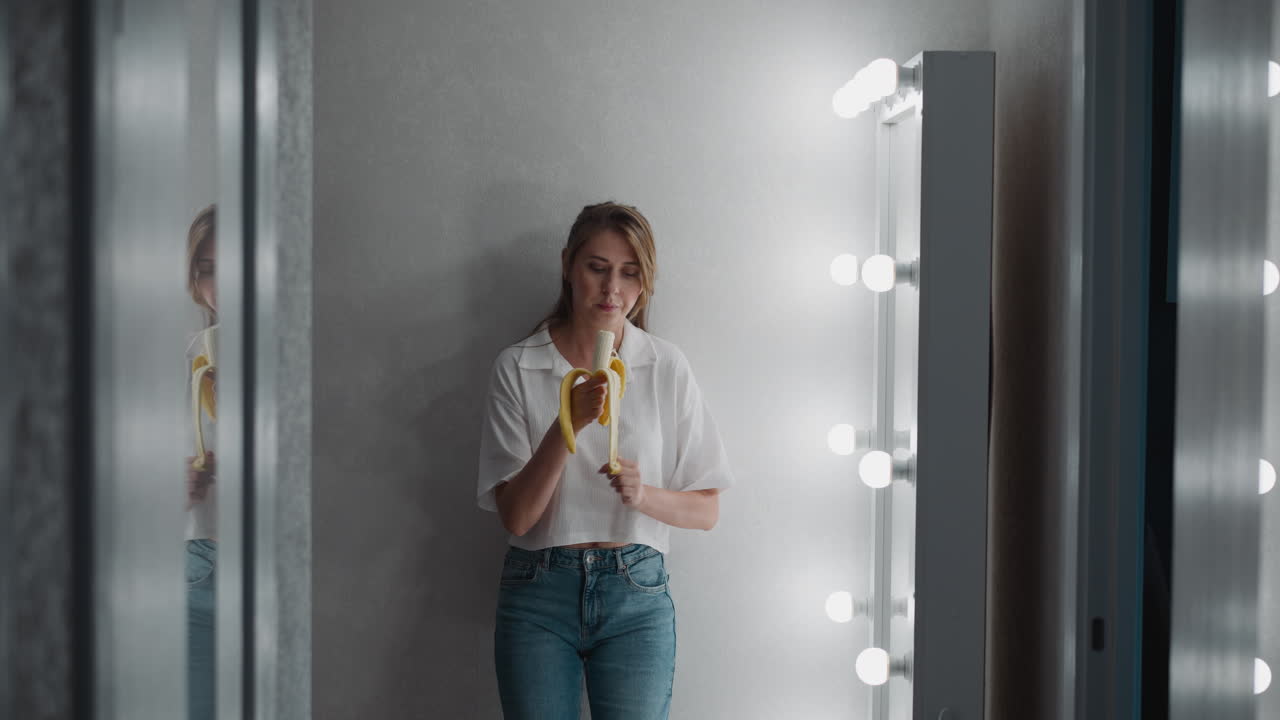 girl in casual wear rests against wall peeling banana and eating it while placing hand inside pocket beside mirror decorated with glowing bulbs in bright relaxed indoor atmosphere