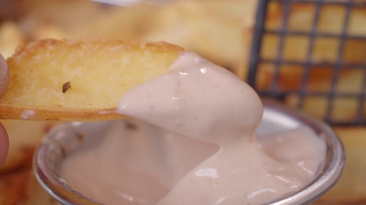 Close up of a hand dipping a crispy potato wedge into a creamy white sauce.