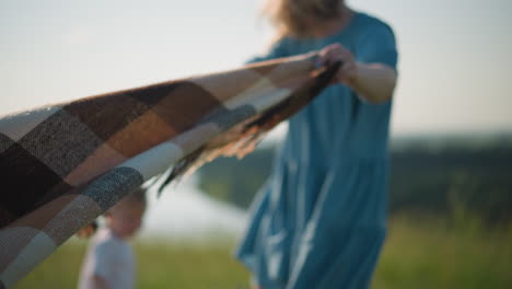 a woman in a blue gown, her face blurred, holds a plaid scarf as the wind gently blows it in a grassy field. her kids are also blurred in the background