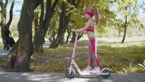 Young-athletic-fit-blonde-girl-rides-traveling-on-electric-scooter-on-road-in-park-on-sunny-day