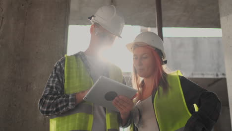 trabajador de la construcción hombre y arquitecto mujer en un casco discutir el plan de construcción de la casa hablar el uno al otro sobre el diseño sosteniendo una tableta mirar los dibujos fondo de los rayos del sol.