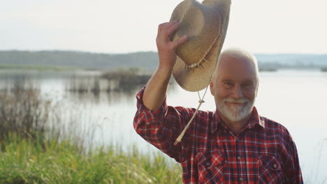 senior man in red shirt standing on the river bank and doing greeting gesture to the camera taking off a hat