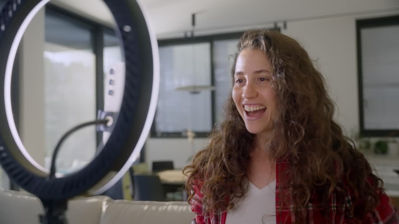 Female influencer or vlogger using ring light and phone to film herself talking on online blog or content channel. Creator on social media happy to speak to fans and followers. Working from her home