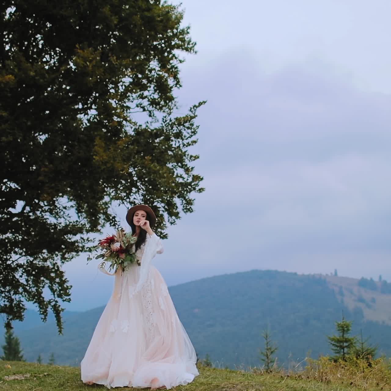 Bride in white dress near the green tree. Beautiful woman in long white dress standing with bouquet of flowers against mountainous area.