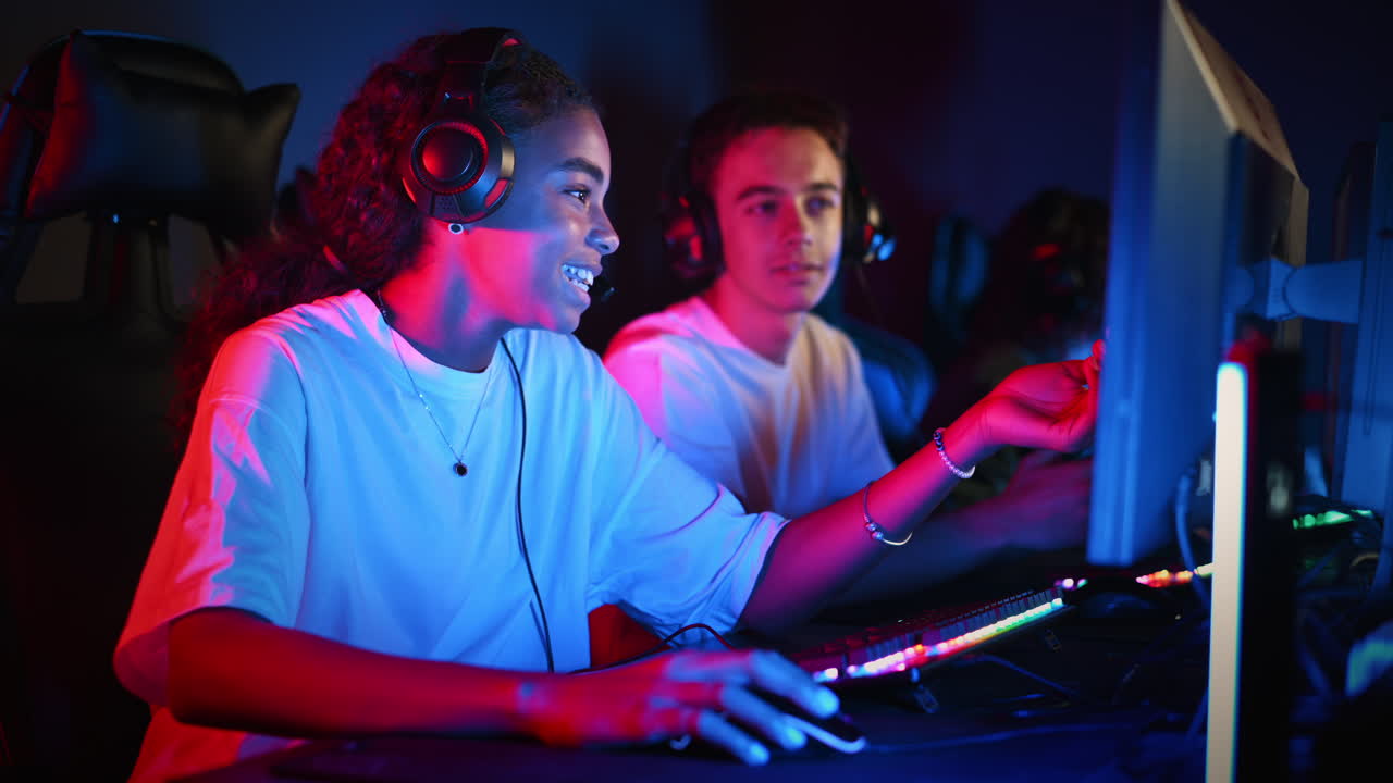 White boy and black girl teens in headsets discussing while playing video games in video game club with blue and red illumination. Keyboard and mouse with illumination